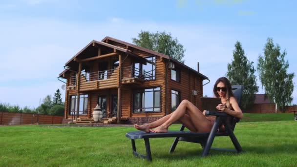 Uma bela mulher esbelta se encontra em um chaise longue em um maiô com um tablet no fundo de uma grande casa de campo em óculos de sol e banhos de sol ao sol visualizando fotos em redes sociais — Vídeo de Stock