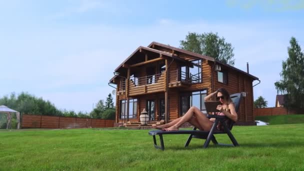 Une belle femme mince se trouve sur une chaise longue dans un maillot de bain avec une tablette sur le fond d'une grande maison de campagne en lunettes de soleil et fait des achats en ligne — Video
