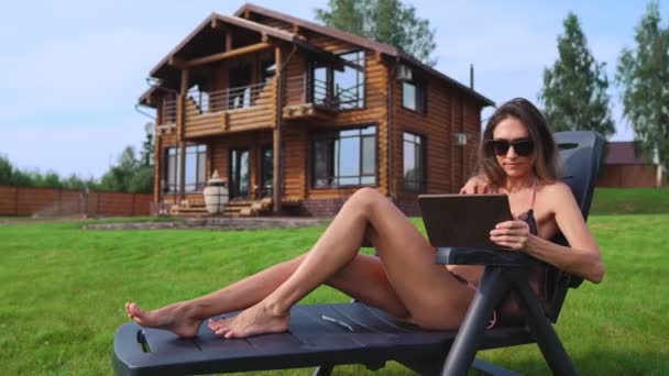 Une belle brune en maillot de bain couchée sur une chaise longue près de son manoir chic avec de grandes fenêtres sourit tout en travaillant à distance à la maison via Internet dans le monde des affaires. Le concept de pays — Video