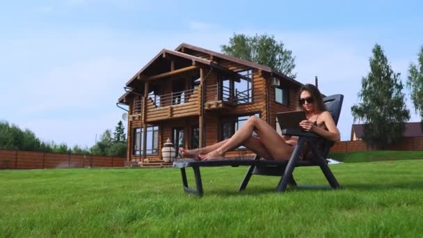 Krásná brunetka v plavkách ležící na lehátku poblíž její elegantní rezidence s velkými úsměvy, Windows a vlny ji za ruku k přátelům a práce vzdáleně na tabletovém počítači — Stock video