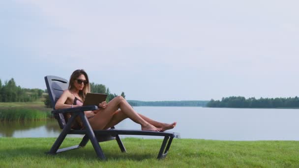 Nő feküdt egy nyugágyon a fürdőruhát, egy tabletta számítógép finger megérinti a képernyőt, a tabletta és a mosoly, a tó partján fekvő — Stock videók