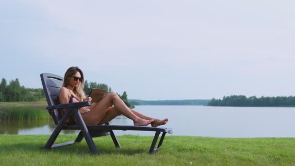 Mooie brunette in een zwembroek liggend op een ligstoel op de meer glimlachen werken op afstand via het Internet in de zakelijke sfeer. Het concept van het landelijke leven — Stockvideo