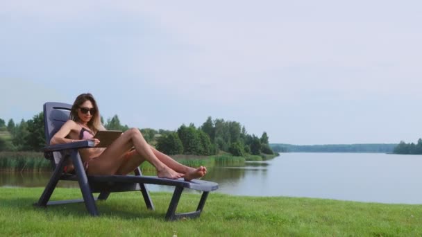 Mooie brunette in een zwembroek liggend op een ligstoel op de meer glimlachen werken op afstand via het Internet in de zakelijke sfeer. Het concept van het landelijke leven — Stockvideo