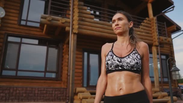 黑色顶部有漂亮腹肌的运动型黑发妇女在锻炼后, 在木制豪宅的背景下出汗, 喝一瓶水 — 图库视频影像