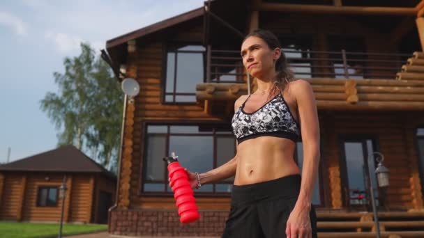 Crooked brunette with inflated press muscles and black shorts, drinking protein after a workout on the street against the background of a huge house with large Windows — Stock Video