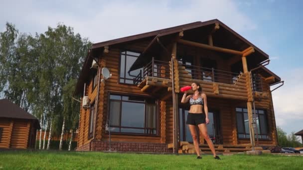 スポーティなブルネット女性黒トップの素敵な腹筋と木造の大邸宅の背景に汗で濡れて、トレーニングの後は、ボトルから水を飲む — ストック動画