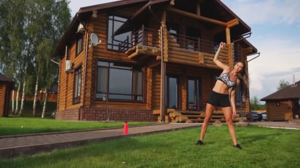 A slim and beautiful woman in sports clothes with an open press is preparing to start training on the lawn near her house Performing slopes flexing back muscles — Stock Video