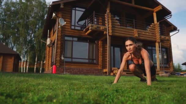 A slender young woman in black sportswear performs squats on the grass near her country house — Stock Video