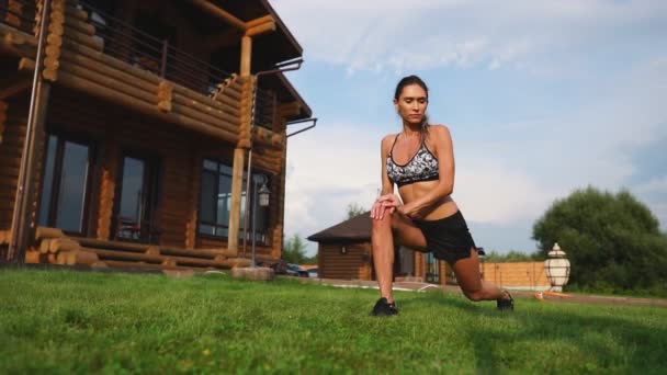 En smal och vacker kvinna i sportkläder med en öppen press förbereder sig för att börja träna på gräsmattan nära sitt hem som flexar sin ben, knän, Smalben och lår med värma upp övningar — Stockvideo
