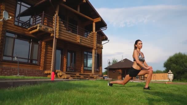 Une femme mince et belle en tenue de sport avec une presse ouverte se prépare à commencer l'entraînement sur la pelouse près de sa maison en fléchissant ses jambes, genoux, tibias et cuisses avec des exercices de réchauffement — Video