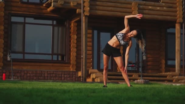 Una mujer delgada y hermosa en ropa deportiva con una prensa abierta se prepara para comenzar a entrenar en el césped cerca de su casa Realizar pendientes flexionando los músculos de la espalda — Vídeo de stock