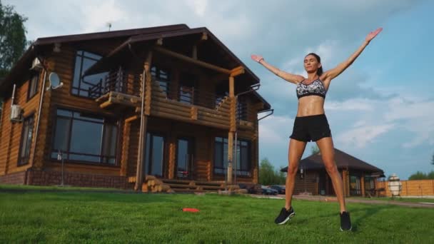 Slim krásná žena v oblečení na pozadí domu na trávníku provádí skoky pro kardio trénink a spalování tuků — Stock video