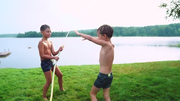 Junge in Badehose gießt Wasser auf seinen jüngeren Bruder, der sich im Park auf der Wiese am See vergnügt — Stockvideo