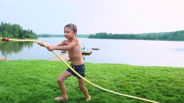 Junge in Badehose gießt Wasser auf seinen jüngeren Bruder, der sich im Park auf der Wiese am See vergnügt — Stockvideo