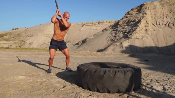 Muscle athlète homme fort homme frappe un marteau sur une énorme roue dans les montagnes sablonneuses au ralenti au coucher du soleil. La poussière des roues monte . — Video