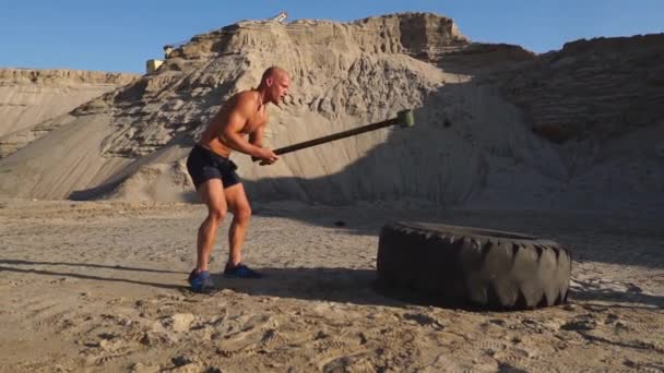 Muscle athlète homme fort homme frappe un marteau sur une énorme roue dans les montagnes sablonneuses au ralenti au coucher du soleil. La poussière des roues monte . — Video