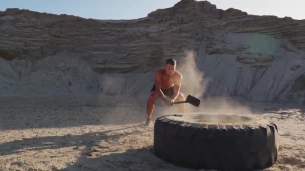 Atleta muscular homem forte bate um martelo em uma roda enorme nas montanhas arenosas em câmera lenta ao pôr do sol. A poeira das rodas sobe . — Vídeo de Stock