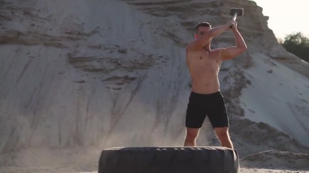 Сильный спортсмен бьет молотком по огромному колесу в песчаных горах в замедленной съемке на закате. Пыль с колес поднимается . — стоковое видео