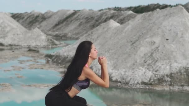 Deportiva chica morena en ropa deportiva negro entrena contra una cantera de arena de tiza blanca con agua azul. Saltos en cada pierna para los muslos — Vídeo de stock