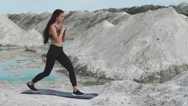 Deportiva chica morena en ropa deportiva negro entrena contra una cantera de arena de tiza blanca con agua azul. Saltos en cada pierna para los muslos — Vídeos de Stock