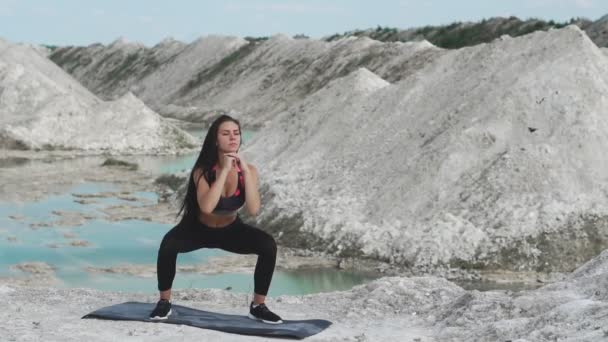 Sportieve brunette meisje in zwarte sportkleding treinen tegen een steengroeve wit krijt zand met blauwe water. Uitvoeren van squats — Stockvideo