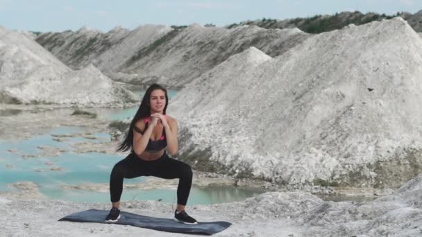 Ragazza bruna sportiva in abbigliamento sportivo nero si allena contro una cava di sabbia bianca di gesso con acqua blu. Esecuzione di squat — Video Stock