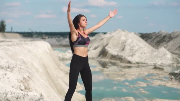 Deportiva chica morena en ropa deportiva negro entrena contra una cantera de arena de tiza blanca con agua azul. Realización de saltos de burpee en el acantilado . — Vídeos de Stock