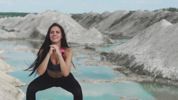 Fille brune sportive en tenue de sport noire s'entraîne contre une carrière de sable blanc à la craie avec de l'eau bleue. Exécution de squats pour renforcer les muscles des fesses — Video