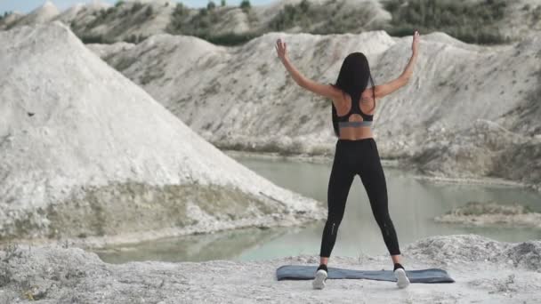Sporty brunette girl in black sportswear trains against a white chalk sand quarry with blue water. Performing burpee jumps on the cliff. — Stock Video