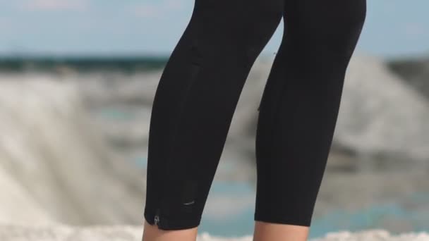 Menina morena esportiva em trem sportswear preto contra uma pedreira de areia de giz branco com água azul. Realizando saltos burpee no penhasco . — Vídeo de Stock