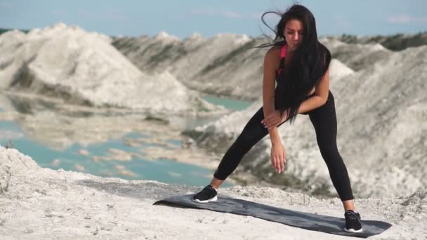 Sportig brunett tjej i svart sportkläder tåg mot en vit krita sand stenbrottet med blå vatten. Utför rullar från fot till fot i squat position — Stockvideo