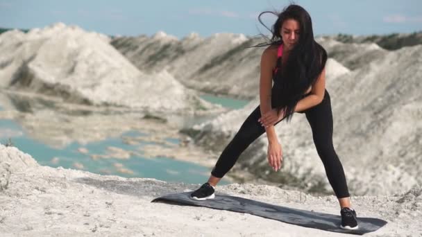 Ragazza bruna sportiva in abbigliamento sportivo nero si allena contro una cava di sabbia bianca di gesso con acqua blu. Esecuzione di rulli da piede a piede in posizione tozza — Video Stock