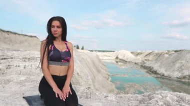 Siyah spor giyim trenler mavi su ile beyaz tebeşir kum ocağı karşı sportif esmer kız. Dağın tepesinde yoga yapmak ve nerede mavi nehir akar mesafe içine bak