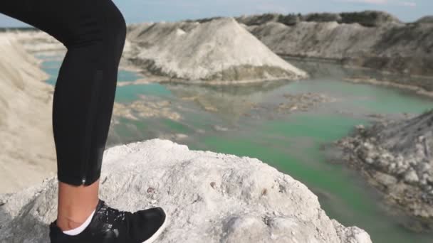 Menina morena esportiva em trem sportswear preto contra uma pedreira de areia de giz branco com água azul. Close-up de pés em tênis se encaixam na borda da montanha com vista para o rio azul . — Vídeo de Stock