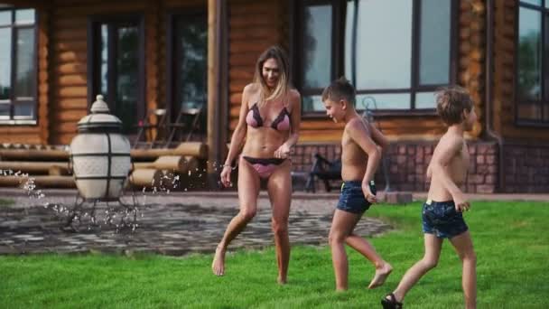 Lycklig familj med två barn som leker med trädgård slang hälla vatten på gräs — Stockvideo