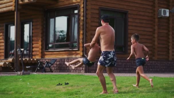 Papa cirkelen zijn zoon in zijn armen zoals op de carrousel, en naast de moeder en de tweede zoon spelen met een slang voor drenken en dousing elkaar met water — Stockvideo