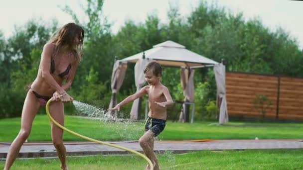 Мама і син, граючи на газон розливу води сміятися і весело провести час на ігровому майданчику з газон на тлі його будинку біля озера — стокове відео