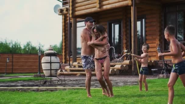 Mère et père jouant avec les enfants sur la pelouse de la maison versant de l'eau d'un tuyau — Video