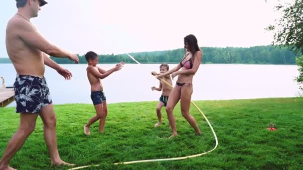 Μητέρα με τον πατέρα και δύο παιδιά που παίζουν στην του γκαζόν ρίχνει νερό γελώντας και διασκεδάζοντας στην παιδική χαρά με γρασίδι στο φόντο του σπιτιού του, κοντά στη λίμνη σε αργή κίνηση — Αρχείο Βίντεο