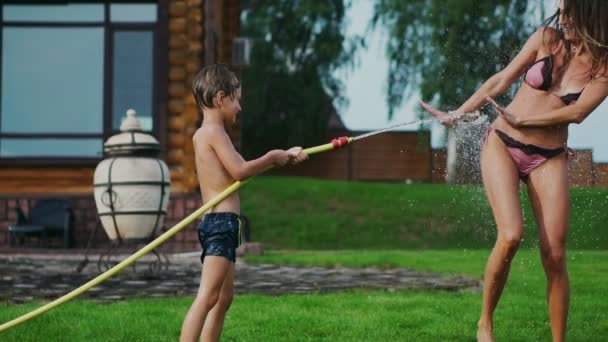 Mamá papá y dos hijos en el verano en la hierba vertían agua de una manguera para el césped. Casa de campo cerca de un lago al atardecer — Vídeos de Stock