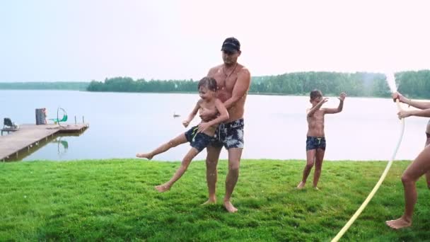 Mãe pai e dois filhos no verão na grama derramou água de uma mangueira para o gramado. Área casa de campo perto de um lago ao pôr do sol — Vídeo de Stock