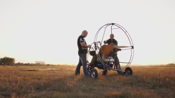 Motorgleitschirm steht bei Sonnenuntergang mit Holzpropeller auf einem Feld, zwei Piloten wärmen den Motor vor dem Flug auf. ein Testlauf aller Systeme — Stockvideo