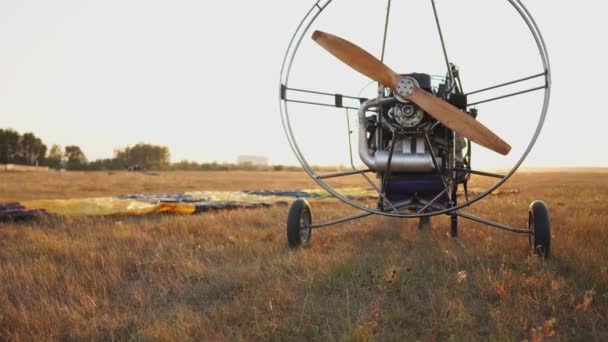 Der Motorgleitschirm steht bei Sonnenuntergang mit einem hölzernen Propeller auf dem Feld, und der Pilot legt den Fallschirm aus und richtet die Schlingen aus — Stockvideo