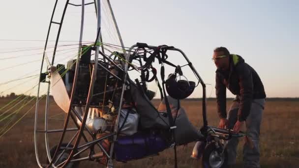 Um homem piloto de um parapente de motor prende o paraquedas ao corpo do parapente preparando-se para o voo e verificando o equipamento — Vídeo de Stock