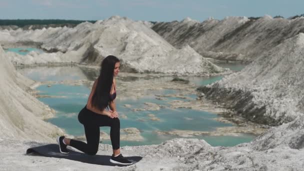 Sportowy brunetka dziewczyna w czarnym sportowej pociągi przeciwko kamieniołom kreda biały piasek z błękitną wodą. Rzuca na każdą nogę na udach — Wideo stockowe