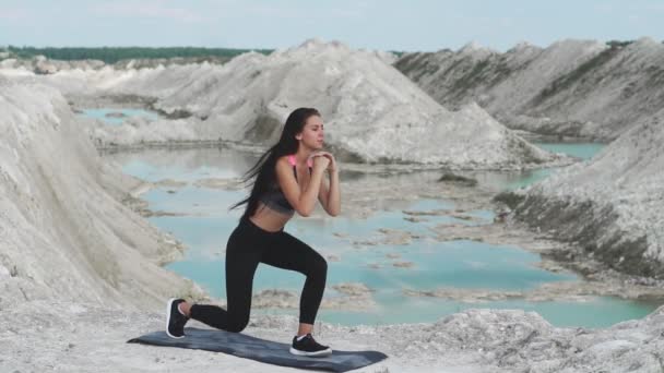 Deportiva chica morena en ropa deportiva negro entrena contra una cantera de arena de tiza blanca con agua azul. Saltos en cada pierna para los muslos — Vídeo de stock