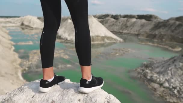 Sportliches brünettes Mädchen in schwarzer Sportbekleidung trainiert gegen einen weißen Kreidesandsteinbruch mit blauem Wasser. Nahaufnahme der Füße in Turnschuhen, die an den Rand des Berges mit Blick auf den blauen Fluss passen — Stockvideo