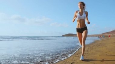 Spor şort ve tişört sahilde yavaş çekimde adada beyaz kumları ve mavi okyanus su ile çalışan güzel bir kadın. Dalgalar ve geri kazanılan kum tepelerinde