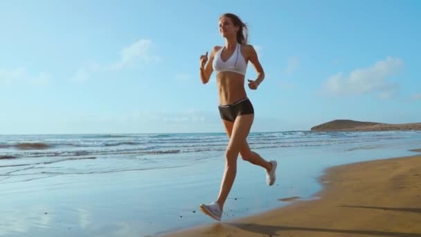 Спортивная женщина бегает по красивому песчаному пляжу, ведет здоровый образ жизни, наслаждается активным летним отдыхом у моря. SLOW MIIION STEADICAM . — стоковое видео
