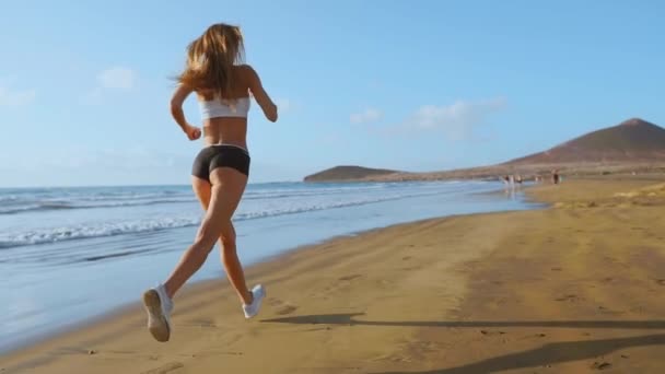 Спортивные женщины в белых майках, бегущие по красивому песчаному пляжу, здоровый образ жизни. Вид сзади. SLOW MIIION STEADICAM . — стоковое видео